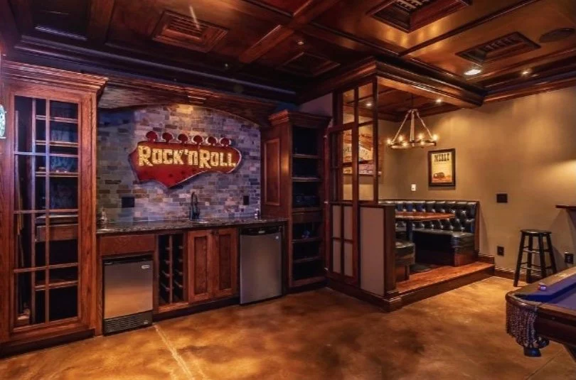 Home bar area with a Rock 'n' Roll sign, wooden cabinets, mini fridge, wine rack, and a seating nook with a black leather bench and wall art.