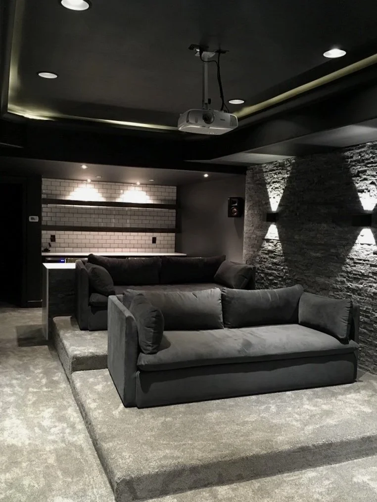 Modern basement home theater with dark gray couches on a raised gray carpeted platform, white brick accent wall, stone textured wall with wall-mounted lights, ceiling mounted projector, and built-in sound speakers.