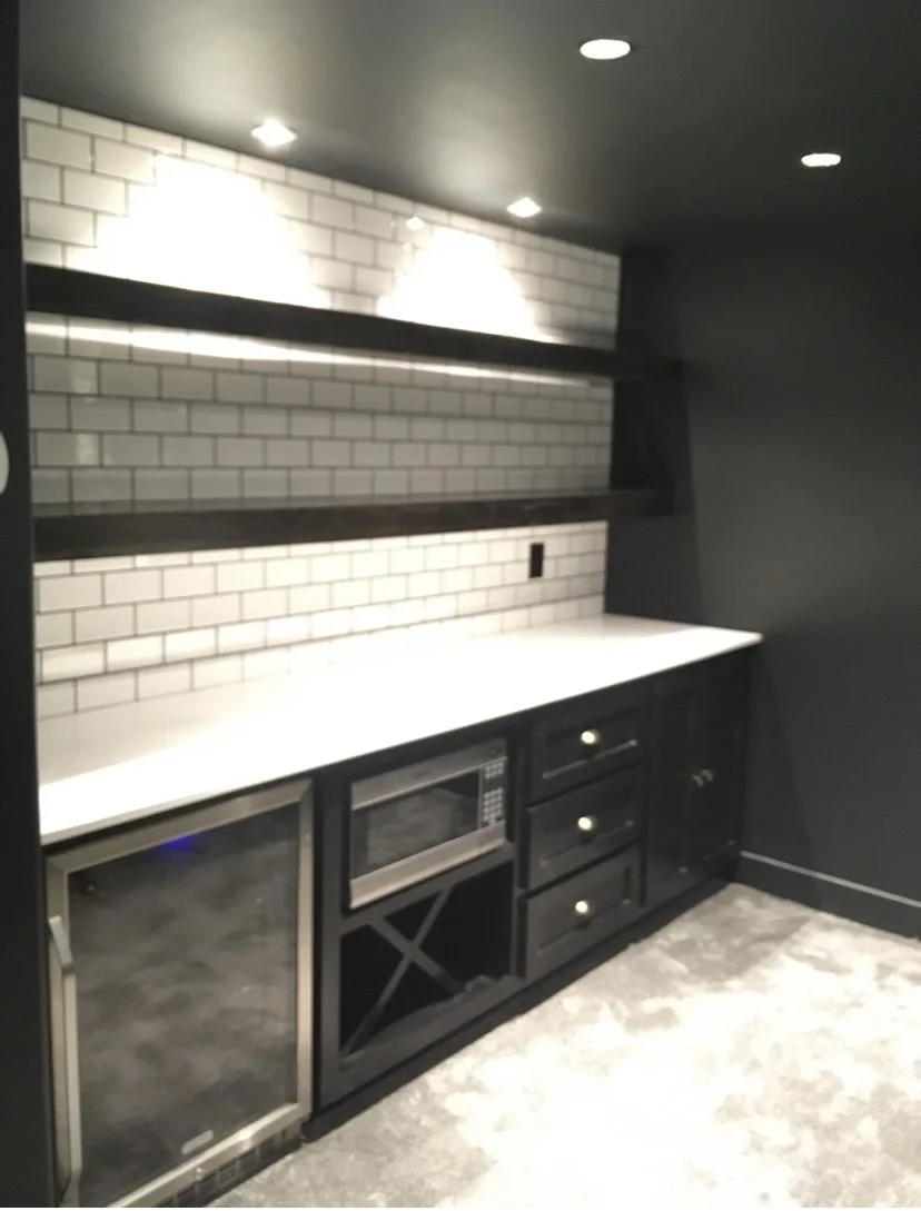 A small bar or kitchenette area with black cabinetry, white countertop, white subway tile backsplash, and open black shelves, integrated microwave, mini fridge, and gray carpeting.
