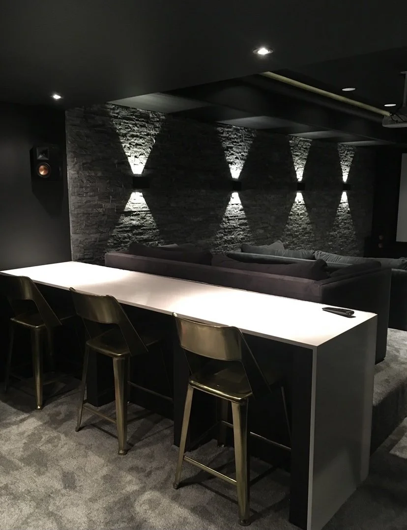 Modern lounge area with a high white counter, four gold chairs, a black stone wall with four wall-mounted lights creating diamond-shaped shadows, and gray couches in the background.