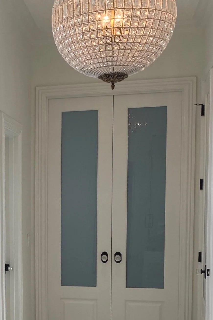 A white double door with four glass panels, surrounded by white trim, with a crystal chandelier hanging from the ceiling above.