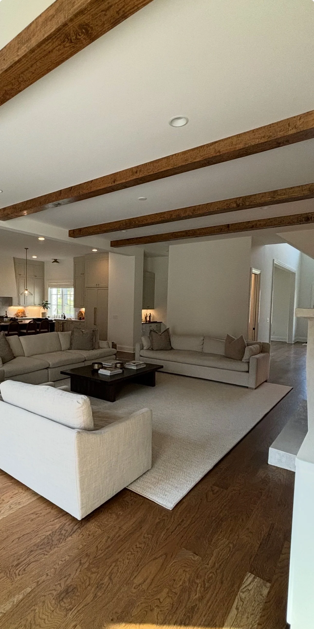 Living room with beige sofas, a black coffee table, wooden floor, white walls, and exposed wooden ceiling beams.