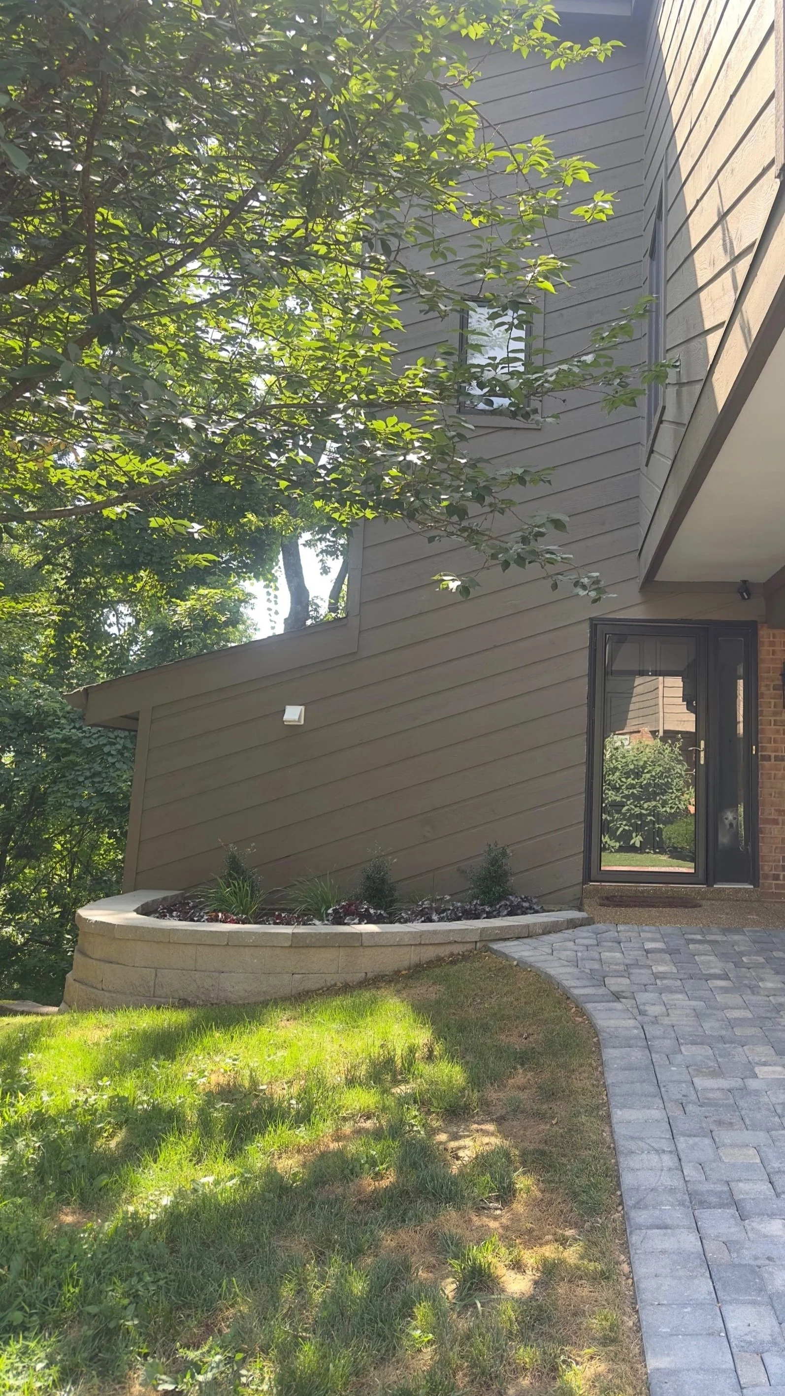 Side view of a house with a dark gray exterior, a front door with a glass panel, a curved stone walkway, a small landscaped garden bed with plants, and a large leafy tree providing shade.