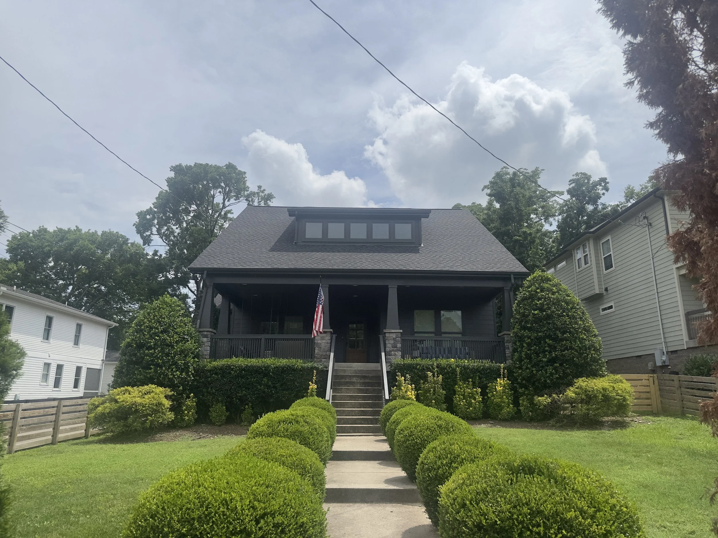 A dark-colored house with a front porch and stairs, surrounded by neatly trimmed bushes and green lawn, with neighboring houses and tall trees in the background under a partly cloudy sky.